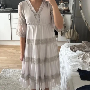Vit klänning - Romantisk vit klänning med v-ringning och spetsdetaljer längs halsringning, ärmar och kjol. Klänningen har trekvartsärmar i transparent tyg och flera lager med spetsband. Den har en luftig, flowy passform och faller strax under knäet.