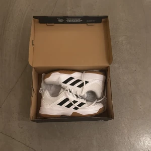 Adidas vita sneakers strl 38 - Fräscha vita Adidas sneakers med klassiska svarta ränder och gummisula i beige. Skorna har snörning och en platt sula med mönstrad undersida för extra grepp. Ovandelen är i syntetmaterial och mesh för bra ventilation. Perfekt för dig som gillar sportig stil.