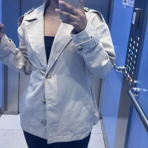 Beige trenchcoat med bälte - Säljer en beige trenchcoat med klassisk krage, knappar framtill och bälte i midjan. 