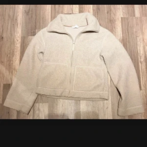 Beige teddyjacka med dragkedja - Mysig beige teddyjacka med hög krage och två stora fickor framtill. Jackan har hel dragkedja och är tillverkad i fluffigt teddy-material som håller dig varm och skön. Perfekt för kyliga dagar och enkel att matcha med olika outfits.