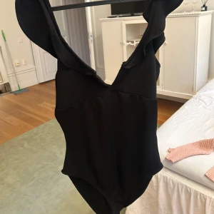 Svart body med volangdetalj - Supersnygg svart  body med djup v-ringning och breda axelband med volangdetaljer. Perfekt för  en stilren och trendig look. Enkel men ändå elegant design som passar dig som vill sticka ut lite extra.