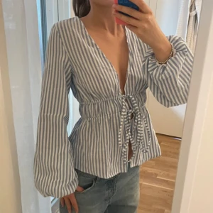 Blåvit randig blus med snörning - Säljer en blåvit blus med vertikala ränder och djup v-ringning. Blusen har långa, lite puffiga ärmar och snörning framtill i midjan som ger en snygg siluett. Perfekt för dig som gillar en chill och trendig look.