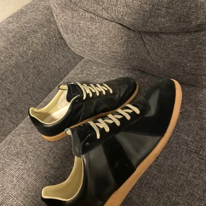 Maison Margiela gats - Maison Margiela sneakers i svart skinn och mocka med beige sula och vita snören. Klassisk låg modell med rund tå och diskret branding på plösen och sulan. Insidan är fodrad i ljusbeige och skon har en stilren, minimalistisk look. Ett par väldigt snygga skor.