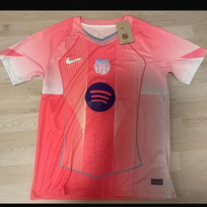 FC Barcelona x Nike rosa fotbollströja M - Snygg FC Barcelona fotbollströja från Nike i rosa och ljusröd med vita detaljer. Tröjan har korta ärmar, Dri-FIT-material och tryckta loggor från både Nike och Spotify. Unik design med tunna linjer och klubbmärke på bröstet. Perfekt för dig som älskar fotboll och vill sticka ut.