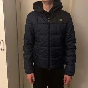 Mörkblå pufferjacka från Lacoste - Säljer en mörkblå pufferjacka från Lacoste med svart axelparti och huva. Jackan har quiltad design, dragkedja framtill och långärmade ärmar. Klassisk Lacoste-logga på bröstet. Perfekt för kalla dagar och har en clean, sportig look. Han på bilden är 175.