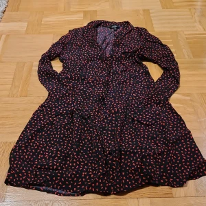 Svart klänning med röda hjärtan & Other Stories - Supersöt svart klänning från & Other Stories med röda hjärtformade mönster. Klänningen har lång ärm, klassisk krage och knappar framtill. Den är kort i modellen och har en flowy passform som känns både lekfull och trendig.