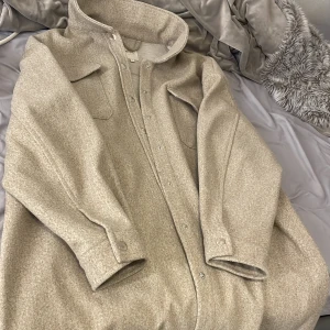 Beige overshirt jacka från ONLY - Säljer en beige overshirt jacka från ONLY i storlek L. Jackan har två stora bröstfickor med lock, knappar hela vägen och en mjuk ullblandad yta. Perfekt för lager-på-lager och har en clean, loose passform med klassisk krage.