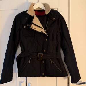 Svart quiltad jacka från Barbour - Quiltad svart jacka från Barbour med mysig beige teddyfoder i kragen, bälte i midjan och snygga detaljer som dragkedjor och knappar. Jackan har lång ärm och en cool, figurnära passform som ger en schysst siluett. Perfekt för dig som gillar stil och komfort.