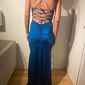 Blå långklänning med snörning i ryggen - Snygg blå långklänning med djup v-ringning och smala axelband som korsas och snöras i ryggen. Klänningen har figurnära passform upptill och faller snyggt längs kroppen. Materialet är glansigt och känns lyxigt, perfekt för en statement-look. Den är endast andvänd 1 gång och är i jätte bra skick!
