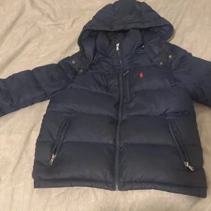 Mörkblå dunjacka från Polo Ralph Lauren - Snygg mörkblå dunjacka från Polo Ralph Lauren med huva och klassisk röd logga på bröstet. Jackan har flera dragkedjefickor, quiltad design och långa ärmar. Perfekt för kalla dagar och har en clean, sportig look.