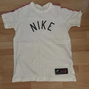 Vit Nike t-shirt med röd detalj - Snygg vit t-shirt från Nike med svart logga framtill och coola röda och svarta ränder längs axlarna. Klassisk passform och korta ärmar. Materialet är mjuk bomull och nere på framsidan finns en patch med Nike Air. Perfekt för dig som gillar sportig streetstyle. Bröst 96cm. Storlek 13-15 år/158-170cm, barn xl storlek men det kan passa vuxen s/m.
