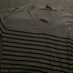 Randig beige/vit v-ringad tröja - Snygg beige tröja med svarta horisontella ränder och v-ringad hals. Tröjan har långa ärmar och är tillverkad i mjukt bomullsmaterial. Perfekt för dig som gillar klassisk stil med en modern twist.