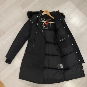  Moose Knuckles damparkas - Svart dunjacka från Moose Knuckles med fluffig päls på huvan, långa ärmar och snygga silverfärgade detaljer. Jackan har dragkedja och knappar framtill, två fickor och quiltad insida. Perfekt för kalla dagar med stil.