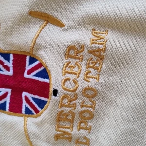 Gul och marinblå pikétröja Ralph Lauren - Snygg pikétröja från Ralph Lauren i gul och marinblå med broderad Union Jack, Mercer Polo Team-text och stor polospelare på bröstet. Tröjan har korta ärmar, klassisk krage med knappar och stor siffra 3 på ryggen. Perfekt för en sportig och preppy stil.