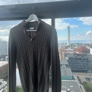 Grå kabelstickad tröja John Henric - Kabelstickad grå tröja från John Henric med halv dragkedja och hög krage. Tröjan har lång ärm och är tillverkad i mjukt stickat material, perfekt för kyligare dagar. Klassisk och stilren design som funkar till många outfits. Pris i butik är 899kr