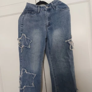 Blå jeans med stjärnformade slitningar - ⚠️ Skriv till mig innan köp!⚠️Säljer ett par blå jeans med unika stjärnformade slitningar på benen. Jeansen har klassisk femficksdesign, raka ben och är tillverkade i denim. Perfekta för dig som vill sticka ut med en cool och trendig look.
