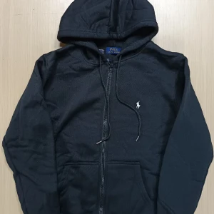 Svart hoodie från Polo Ralph Lauren - Svart hoodie från Polo Ralph Lauren med dragkedja och huva. Klassisk design med liten vit logga på bröstet och snörning vid huvan. Tillverkad i mjuk bomullsmix, perfekt för chill dagar. Fickor framtill och ribbade muddar för extra komfort. Den passar personer mellan 1,70 till 1,80