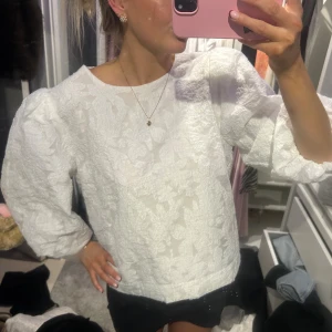 Neo noir blus - Säljer en vit blus med puffiga trekvartsärmar och snyggt strukturerat tyg med blommigt mönster. Blusen har rund hals och en loose passform som ger en chill och trendig vibe. Perfekt att styla med jeans eller kjol för en cool look.