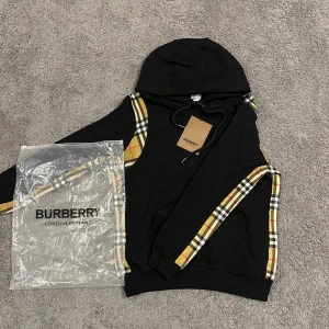 Svart Burberry hoodie med rutiga detaljer - Snygg svart hoodie från Burberry med klassiska rutiga paneler i beige, rött och svart längs ärmarna och axlarna. Tröjan har huva med snörning och känguruficka framtill. Perfekt för dig som vill ha en streetig och lyxig vibe.