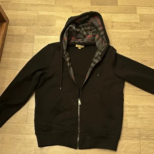 Svart Burberry hoodie med rutig huva - Svart hoodie från Burberry i storlek M med dragkedja och klassisk rutig huva i grått och rött. Tröjan har fickor framtill och snörning i huvan. Perfekt för dig som vill ha en stilren och lyxig streetwear-look.