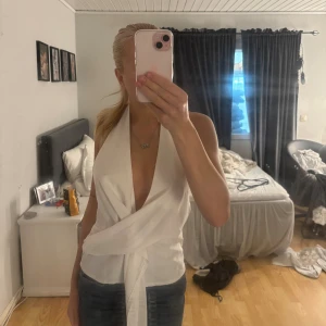 Vit omlott halterneck topp - Säljer en vit omlott halterneck topp med djup v-ringning och öppen rygg. Toppen är ärmlös och har knytning i midjan för en snygg passform. Perfekt för dig som gillar stilrena och trendiga plagg med lite edge.