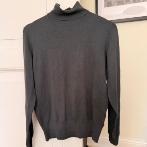 Grå polotröja från H&M - Mörkgrå polotröja från H&M i mjuk och tunn stickad bomullsmix. Klassisk modell med lång ärm och ribbad krage som sitter snyggt runt halsen. Perfekt att styla med jeans eller kjol för en clean look.