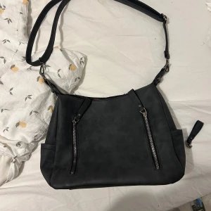 Mörkgrå axelväska med dragkedjor - Säljer en mörkgrå axelväska i syntetmaterial med två snygga dragkedjor framtill och en extra ficka med dragkedja på baksidan. Väskan har justerbar axelrem och flera fack för att hålla ordning på dina grejer. Kedjan där uppe krångla som man kan se så klippt bort den men själva den man stänger med är kvar så de matchar, det går ju att sy en ny dragkedja - jag kan göra det åt dig innan jag skickar men då kostar den extra (samma pris som kjendjan kosta)💕