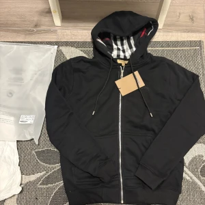 Svart hoodie från Burberry med dragkedja - Svart hoodie från Burberry med klassiskt rutigt foder i huvan och broderad logga på bröstet. Tröjan har hel dragkedja, snörning i huvan och två fickor framtill. Perfekt för dig som gillar stilrena och exklusiva plagg.