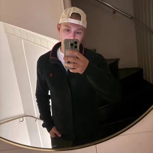 Mörkblå stickad kofta från Polo Ralph Lauren - Säljer en mörkblå stickad kofta från Polo Ralph Lauren med dragkedja och röd logga på bröstet. Koftan har långärmad passform, ribbade muddar och en snygg röd detalj längs kragen. Perfekt för lager-på-lager under höst och vinter.