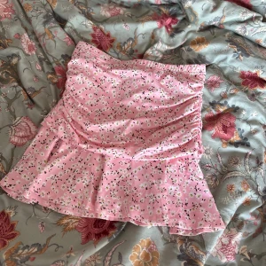 Rosa blommig volangkjol - Supersöt rosa kortkjol med små vita blommor, volangkant och rynkade detaljer på sidorna. Elastisk midja för skön passform. Storlek S🩷