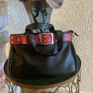 Svart handväska med röd bältdetalj - Cool svart handväska i skinn med en bred röd bältdetalj och silvrigt spänne runt öppningen. Väskan har två rejäla handtag och stängs med dragkedja upptill. Perfekt storlek för det viktigaste och ger en edgy vibe till din outfit.