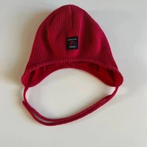 Röd mössa från Polarn O. Pyret  - Röd mössa från Polarn O. Pyret med öronlappar och knytband under hakan. Tillverkad i 100% bomull och har en svart logotyp framtill. Perfekt passform för små barn och mjuk ribbad struktur för extra komfort.