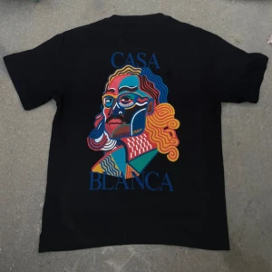 Svart Casa Blanca t-shirt med färggrafik - Svart t-shirt från Casa Blanca med en stor, färgstark grafisk print på ryggen och mindre print på bröstet. Motivet har vågiga linjer i blått, gult, rött och grönt, och texten 'CASA BLANCA' i blått. Klassisk rund hals och korta ärmar. Perfekt för dig som gillar statement-plagg.