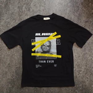 Svart t-shirt med tryck från Engell Z - Cool svart t-shirt från Engell Z med grafiskt tryck framtill i vitt och gult. Motivet har texten 'ALWAYS BETTER THAN EVER' och en bild på en person med solglasögon, samt gula band med text. T-shirten är i mjuk bomullsblandning och har normal passform och korta ärmar.