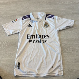 Real Madrid Benzema matchtröja Adidas - Säljer en vit Real Madrid fotbollströja från Adidas med Benzema och nummer 9 på ryggen. Tröjan har korta ärmar, klassisk krage och lila detaljer på axlarna. Materialet är lätt och andas med Climacool-teknologi. Officiell klubbmärke och sponsortryck på bröstet.