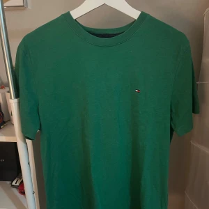 Grön t-shirt från Tommy Hilfiger - Snygg grön t-shirt från Tommy Hilfiger i klassisk modell med rund hals och liten broderad logga på bröstet. Tillverkad i mjuk bomull och har en normal passform. Perfekt för en clean och stilren look.