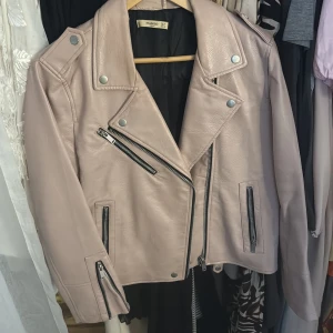 Beige skinnjacka från Mango XL - Säljer en snygg beige skinnjacka från Mango i storlek XL. Jackan har klassisk bikerstil med silvriga dragkedjor, tryckknappar och coola detaljer på axlar och ärmslut. Perfekt för dig som vill ha en stilren och edgy look.