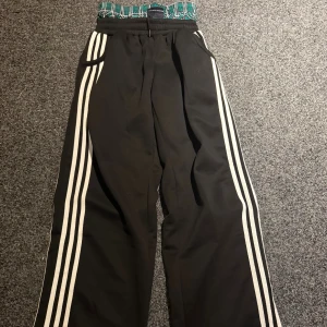 Svarta mjukisbyxor Concrete Mansions - Svarta mjukisbyxor med vita ränder längs benen från Concrete Mansions. Byxorna har elastisk midja med grönt och vitt rutmönster, snörning och sidofickor. Skön loose passform och sportig vibe, perfekta för chill eller streetwear.