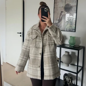 Beige rutig overshirt med fickor - Säljer en beige och grå rutig overshirt med stora bröstfickor och knäppning framtill. Jackan har en avslappnad passform och är tillverkad i ett mjukt, ull-liknande material. Perfekt för lager-på-lager och snygg till både jeans och leggings.