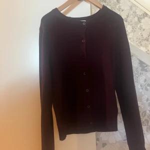 Mörk vinröd kofta med knappar - En stilren vinröd kofta med rund halsringning och knäppning framtill. Koftan har långa ärmar och ribbade muddar vid ärmslut och nederkant. Bara använt någon gång 
