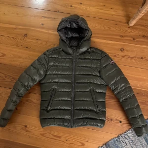 Colmar jacka  - Säljer en mörkgrön pufferjacka från Colmar med huva och dragkedja framtill. Jackan har två fickor med dragkedja och quiltad design. finns ett hål men syns inte då det täcks med armen! Kan gå ner i pris 