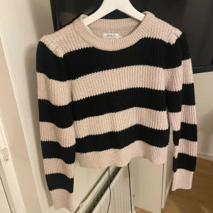 Randig stickad tröja från ONLY - Säljer en snygg stickad tröja från ONLY med breda svarta och beige ränder. Tröjan har en croppad passform, rund halsringning och långa ärmar. Perfekt för dig som gillar en chill och trendig stil.