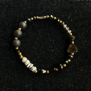 Svart och guldigt pärlarmband med hjärta - Unikt armband med svarta och guldiga pärlor, facetterade detaljer och ett svart hjärta i mitten. Ena sidan har matta svarta pärlor och små randiga pärlor i svart, vitt och grått. Stretchig modell som passar de flesta handleder.