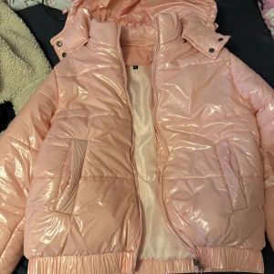Glansig rosa pufferjacka med huva, nypris 600 - Säljer en glansig ljusrosa pufferjacka med huva och dragkedja. Jackan har två stora fickor framtill och resår vid midjan och ärmsluten. Perfekt för kalla dagar och ger en riktigt trendig vibe.