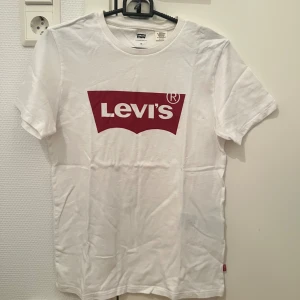 Vit Levi's t-shirt med röd logga XS - Vit t-shirt från Levi's i storlek XS med klassisk röd logotyp framtill. T-shirten har rund halsringning och korta ärmar. Tillverkad i mjuk bomull för en skön och avslappnad känsla. Perfekt för en enkel och stilren look.