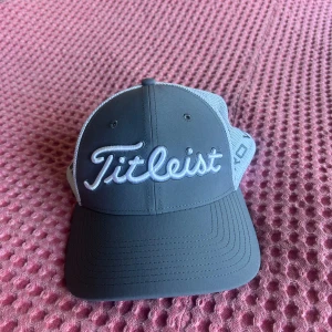 Grå Titleist keps med mesh - Snygg grå keps från Titleist med vit mesh på sidorna och bak. Broderad Titleist-logga framtill, Pro V1 på sidan och FJ på andra sidan. Justerbar snapback baktill med röd och svart Titleist-märke. Perfekt för golf eller sportiga dagar.