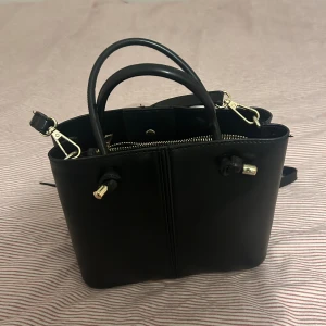 Zara väska  - Snygg svart handväska i skinn med stilrena gulddetaljer och dragkedja. Väskan har både handtag och axelrem, vilket gör den enkel att bära på olika sätt. 