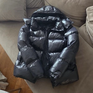 Svart pufferjacka från Everest - Säljer en svart, glansig pufferjacka från Everest med huva och dragkedja. Jackan har ett quiltat mönster och är riktigt fluffig, perfekt för kalla dagar. Märket syns tydligt på ärmen och insidan. Jackan har lång ärm och en loose passform.