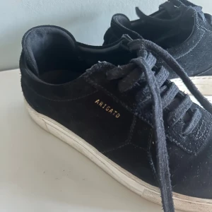 Axel Arigato skor - Snygga svarta sneakers från Axel Arigato i mocka med vit sula och klassisk snörning. Diskret guldtext på sidan och Arigato-logga under sulan. Perfekta för dig som gillar stilrena och trendiga skor med premiumkänsla.