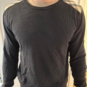 Svart Burberry tröja med rutiga axlar - Svart långärmad tröja från Burberry med klassiska beige och svarta rutiga detaljer på axlarna. Tröjan har rund halsringning och är tillverkad i mjukt material som känns skönt mot huden. Perfekt för dig som gillar stilrena plagg med en twist.